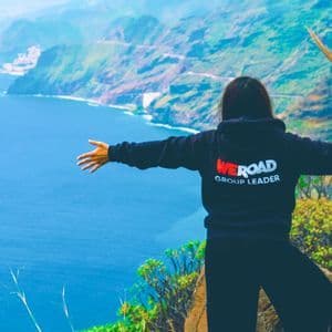 Un capo gruppo WeRoad si erge su una scogliera erbosa con le braccia aperte, ammirando una costa frastagliata e un vasto oceano blu.
