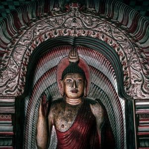 Una statua dipinta di Buddha con la mano alzata, collocata in una nicchia ad arco riccamente decorata.
