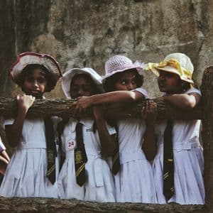 Quattro giovani scolare in uniforme e cappelli guardano oltre una recinzione di legno rustica.