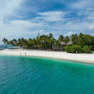 Una vista aerea di una spiaggia tropicale con sabbia bianca, palme e acqua turchese, con due persone che camminano lungo la riva.