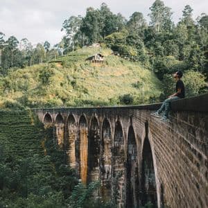 Un uomo in maglietta nera siede sul bordo di un grande ponte ad arco in pietra, guardando una rigogliosa collina verde e boscosa.