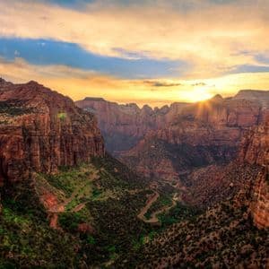 Il sole tramonta su un canyon profondo, illuminando le rosse pareti rocciose e una strada tortuosa nella valle verde sottostante.