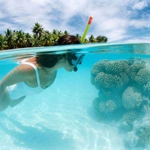 A split-shot view of a woman snorkeling over a coral reef in clear water, with a palm-tree island under a cloudy sky.
