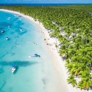 An aerial view of a tropical white sand beach with turquoise water, boats, and a dense forest of palm trees.