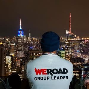 Ein Mann von hinten, der ein 'WeRoad Group Leader'-T-Shirt und eine Beanie trägt und nachts auf eine beleuchtete Stadtkulisse blickt.