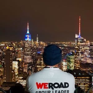 Una persona di spalle, con una maglietta WeRoad Group Leader e un berretto blu, osserva lo skyline illuminato di una città di notte.