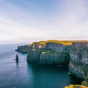 Des falaises escarpées recouvertes d'herbe verte plongent dans un océan bleu calme, avec une aiguille rocheuse à proximité, le tout sous un ciel dégagé.