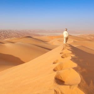 Une personne en tenue traditionnelle marche le long de la crête d'une grande dune de sable dans le désert, laissant des empreintes derrière elle.