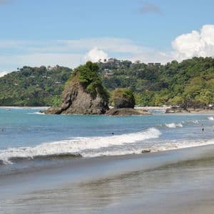 Small waves roll onto a sandy beach where people swim near large rock formations and a lush green hill under a blue sky.
