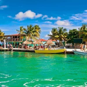 A WeRoad group trip relaxes on a wooden pier by a tropical bar, with boats docked in the turquoise water and palm trees under a blue sky.