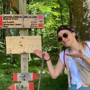 Eine Frau lächelt und zeigt mit einem Stock auf ein Wegweiser-Schild des 'Cammino Kalabria Coast to Coast' im Wald.