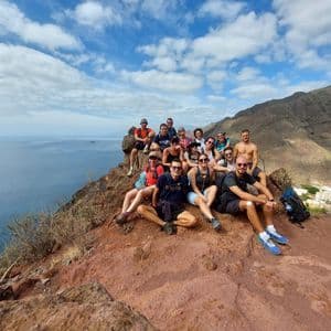 Un gruppo WeRoad in viaggio posa per una foto su una scogliera rocciosa che si affaccia sul mare e sulle montagne sotto un cielo azzurro.