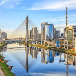 A cable-stayed bridge spans a river in a modern city, with skyscrapers reflected in the calm water under a blue sky.