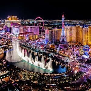 Veduta aerea di uno skyline cittadino di notte, con edifici illuminati, un grande spettacolo di fontane e una replica della Torre Eiffel.