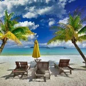 Sdraio vuote e un ombrellone giallo su sabbia bianca tra due palme, di fronte a un oceano turchese sotto un cielo nuvoloso.