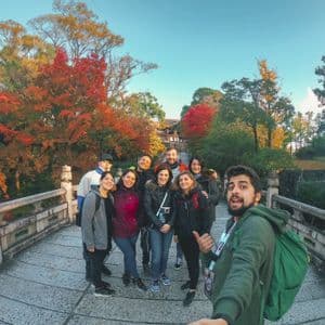 Un viaje en grupo de WeRoad se toma una selfie en un puente de piedra rodeado de árboles con follaje otoñal colorido.