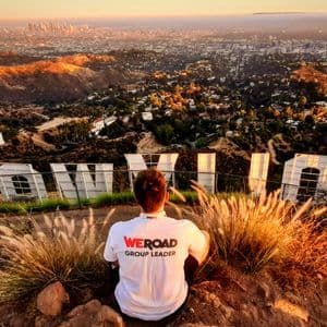 Un Group Leader WeRoad è seduto su una collina dietro la scritta Hollywood, affacciato su una vasta città al tramonto.