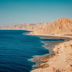 A panoramic view of a deep blue sea meeting a rugged, arid coastline with mountains under a clear sky.