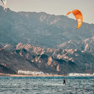 A person kitesurfs across the water with an orange kite, with a large, arid mountain range in the background.