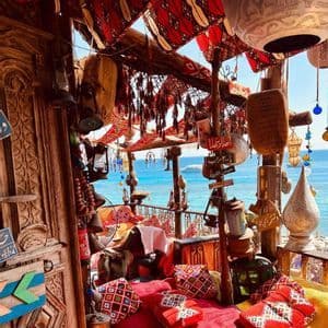 A traditionally decorated balcony with red patterned cushions and hanging lanterns overlooks a bright blue sea on a sunny day.