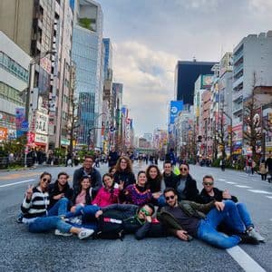 A WeRoad group trip sits together in the middle of a wide city street, posing for a photo with tall buildings in the background.