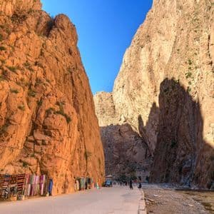 A road lined with market stalls selling colorful textiles winds through a deep canyon with steep, reddish-orange rock walls under a blue sky.