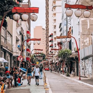 People walk down a busy city street lined with tall buildings and decorated with red, Asian-style lampposts and lanterns.