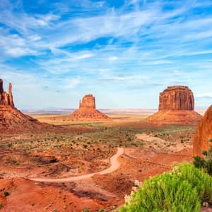Tre grandi butte di arenaria rossa si ergono in una valle desertica, con una strada sterrata tortuosa e arbusti verdi sotto un cielo blu.