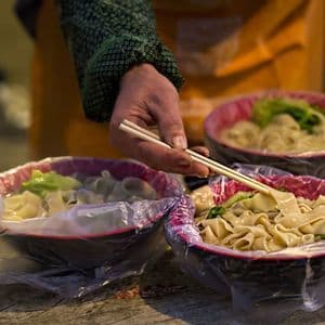 Eine Person serviert an einem Streetfood-Stand mit Essstäbchen flache Nudeln und Gemüse in mit Plastik ausgelegte Schalen.