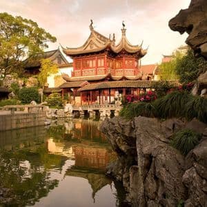 Ein traditioneller roter chinesischer Pavillon mit gestuften Dächern spiegelt sich in einem ruhigen Teich wider, umgeben von üppigen Gärten und Felsen.