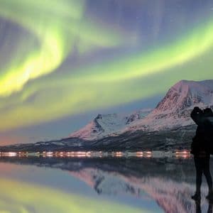 A silhouetted person photographs the green aurora borealis over snow-covered mountains, reflected in calm water at night.
