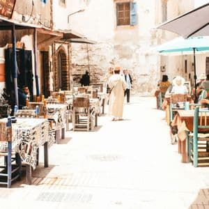 Un caffè all'aperto con tavoli e sedie disposti in una piazza soleggiata e lastricata in pietra, con persone che mangiano e passeggiano.