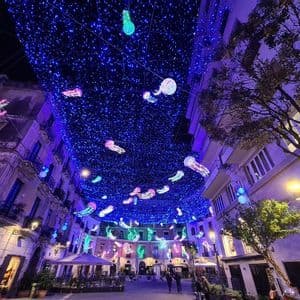 Un soffitto di luci blu e colorate lanterne luminose a forma di medusa pendono sopra una strada cittadina di notte.