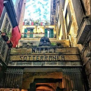 Vista en contrapicado de la entrada de piedra a Napoli Sotterranea, con un rostro tallado sobre el arco en un callejón estrecho.