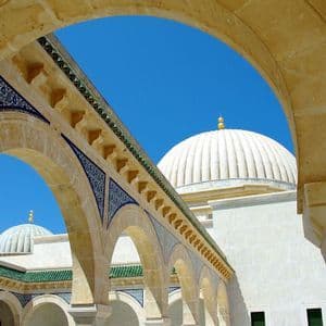 Un arco in pietra color sabbia con dettagli in piastrelle blu incornicia la cupola bianca di una moschea contro un cielo azzurro limpido.