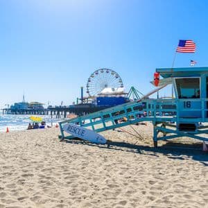 Ein blauer Rettungsschwimmerturm an einem sonnigen Strand, mit einem Pier und einem Riesenrad im Hintergrund unter klarem Himmel.