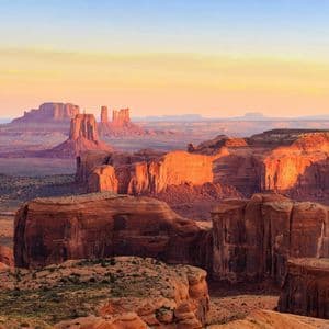 Un vaste paysage désertique avec de grandes formations rocheuses rouges et des buttes illuminées par la lumière chaude d'un soleil levant ou couchant.