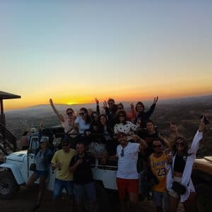 Un gruppo WeRoad in viaggio che posa e festeggia accanto ai fuoristrada su una collina al tramonto.