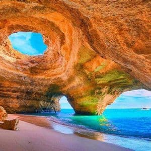 Una spiaggia sabbiosa all'interno di una grotta marina dorata si apre su acque oceaniche turchesi, con un oculo circolare nel soffitto che mostra il cielo blu.