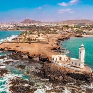 Vista aerea di un faro bianco su una penisola rocciosa vicino a una città costiera con acqua turchese