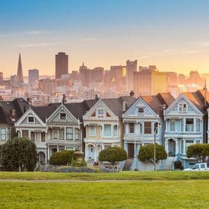 A row of colorful Victorian houses with a modern city skyline in the background during sunset, seen from a grassy park.