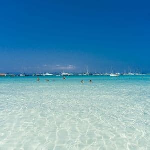 Personas nadando en aguas poco profundas, claras y turquesas con numerosos barcos anclados en el horizonte bajo un vasto cielo azul.
