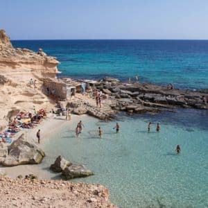 La gente toma el sol en una pequeña playa de arena y nada en las claras aguas turquesas de una cala rocosa en un día soleado.