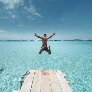 Una persona salta de un muelle de madera a aguas claras y turquesas con los brazos extendidos y barcos al fondo.