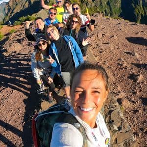 A smiling WeRoad group trip poses for a selfie while hiking along a sunny, narrow mountain ridge.
