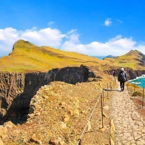 Una persona con una mochila camina por un estrecho sendero de piedra al borde de un acantilado, con vistas a un vasto océano azul bajo un cielo despejado.