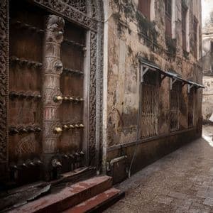 Un homme en vêtement blanc traditionnel marche le long d'une ruelle étroite et humide, passant devant une grande porte en bois richement sculptée.