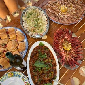 Un pasto all'aperto su un tavolo di legno con vassoi di gamberetti, panini, stufato di verdure e una bottiglia di vino