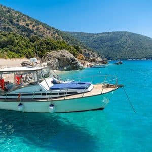 A boat carrying a WeRoad group trip is anchored in clear turquoise water near a hilly coastline with a small beach.