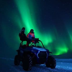 Dos personas con cascos sentadas en un vehículo todoterreno en la nieve, observando la aurora boreal iluminar el cielo nocturno estrellado.